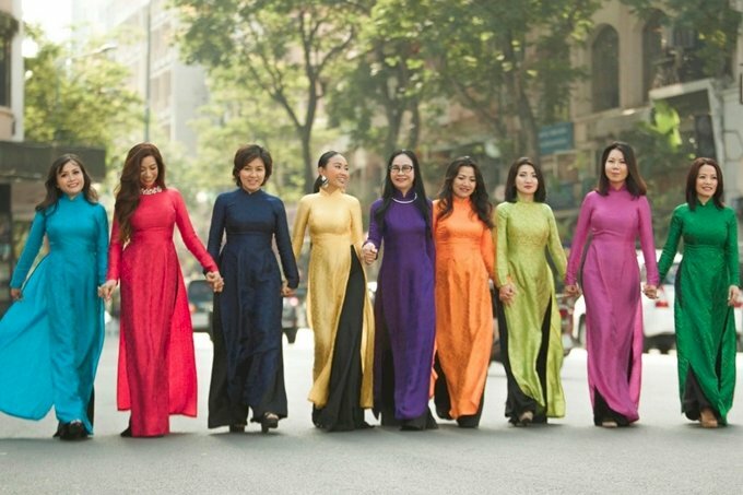 Cách tạo dáng chụp ảnh áo dài tập thể kiểu tự nhiên cho nhóm bạn nữ 9 người, với mỗi người mặc áo dài sắc màu áo dài khác nhau, xếp thành hàng ngang cùng cầm tay nhau bước đi tung tăng dưới phố.