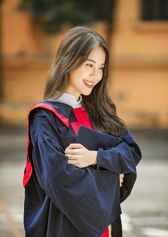 Tạo dáng như thiếu nữ trong bộ áo cử nhân đang cười tươi đầu hơi cúi, 2 tay ôm nón vào lòng