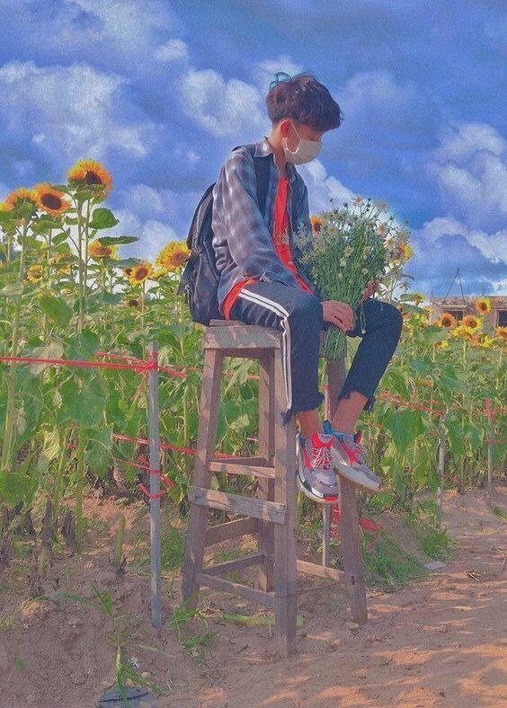 Cách tạo dáng nam học sinh đang ngồi tại cánh đồng hoa hướng dương 2 tay cầm bó hoa dại, mắt nhìn bó hoa dưới khung cảnh trời xanh mây trắng