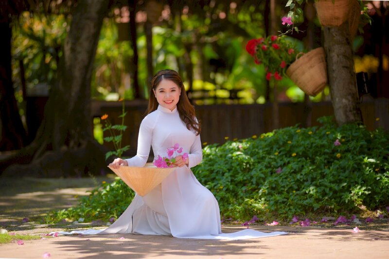 Dáng ngồi duyên dáng khụy gối nghiêng người, 2 tay chạm ành nón lá, một tay cầm nhánh hoa, mặt hướng về ống kính, miệng cười tươi