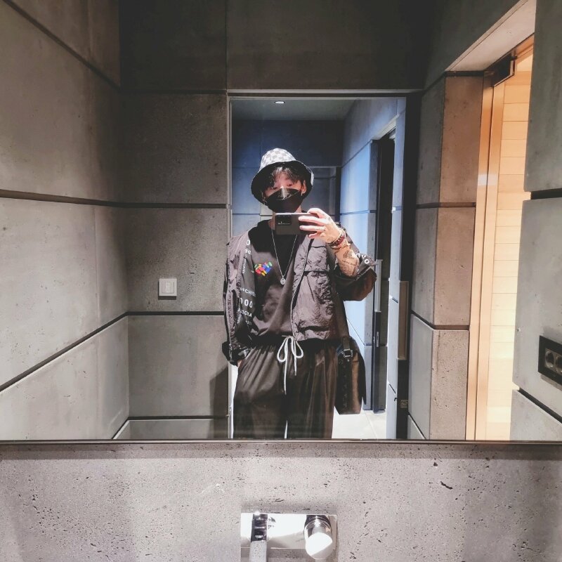 Kiểu selfie nam với bộ quần áo đen cùng chiếc khẩu trang đen, đầu đội nón vành đen cùng những phụ kiện thời trang cá tính, dáng đứng thẳng tự nhiên, một tay bỏ túi quần, tay còn lại tự sướng cùng điện thoại
