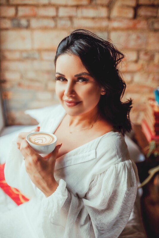 Kiểu tạo dáng cô gái trong chiếc áo trắng xinh đẹp, 2 tay nâng cafe capuchino tạo hình nghệ thuật, mặt hơi ngước, mắt nhìn ồng kính, chụp ở góc nghiêng lấy nửa người