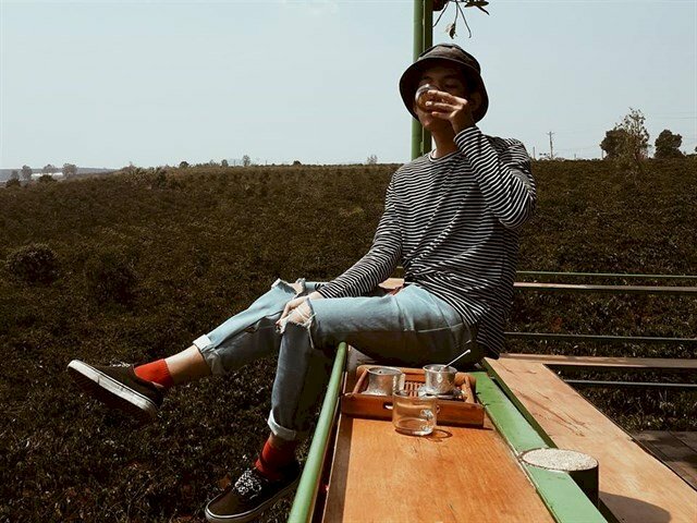 Kiểu tạo dáng nam đang ngồi chill trên coa giữa cánh rừng cao nguyên xanh mướt với 1 tay đặt ở đùi, tay còn lại đang cầm tách cafe uống