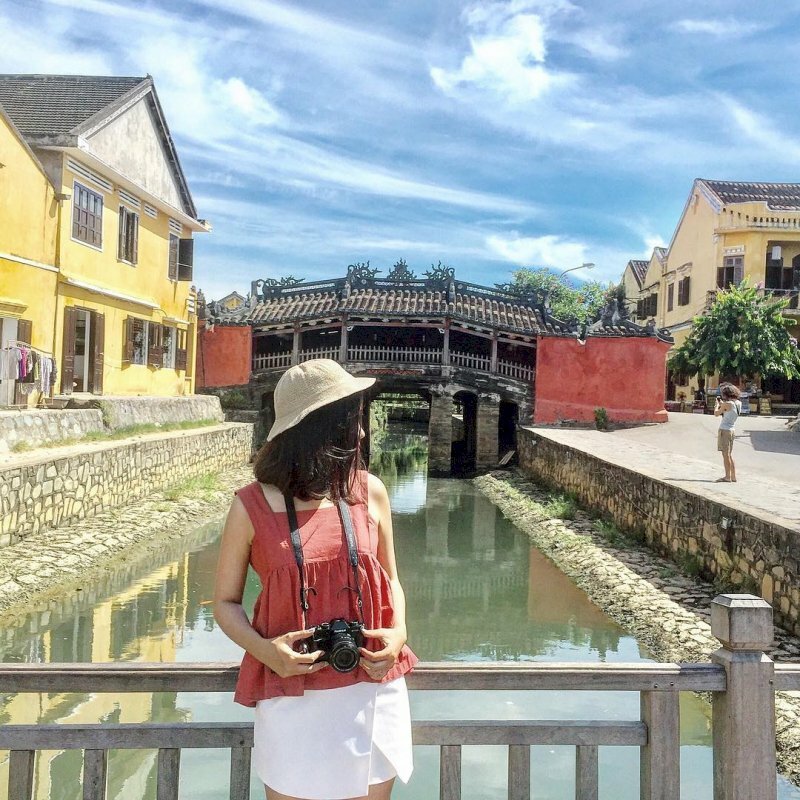 Tạo dáng cô gái đứng tựa lưng vào thành cầu, 2 tay chạm nhẹ vào chiếc máy ảnh cá tính, mặt quay nghiêng chill dưới nắng vàng