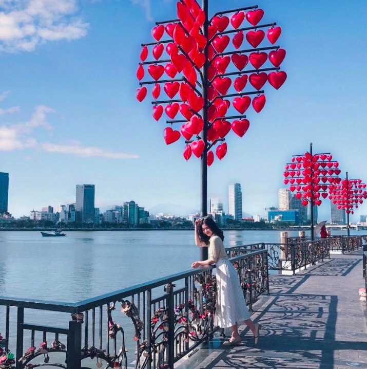 Cách tạo dáng chụp ảnh ở cầu Tình Yêu đẹp lấy trọn 3 cây cột treo đèn lồng trái tim và view sông cực đẹp, với dáng này bạn hãy đứng gần cột đèn lồng và tự bụng vào thành cầu. đứng trụ trên 1 chân, đá nhẹ 1 chân ra sau, 1 tay tựa trên thành cầu và 1 tay đưa lên đầu. 