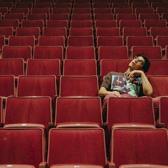 Tạo dáng chụp ảnh trong rạp chiếu phim của chàng trai với cách diển cute trong tư thế ngồi tự nhiên, tay đặt lên đùi, tay còn lại chống càm, đầu hơi nghiêng, mắt nhìn lên trên