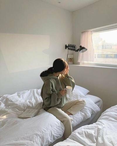 Tạo dáng rất dễ thương của cô gái diện chiếc áo ấm cùng quần kaki đơn giản ngồi co một chân trên giường, một tay cầm cốc cafe, một tay vén tóc, mặt quay ngang hướng ra cửa sổ