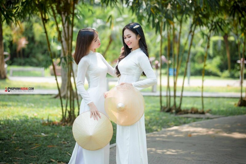 Tạo dáng đẹp cho 2 bạn nữ chơi thân với kiểu đứng nghiêng người vào nhau, tay cầm nón lá, tay còn lại vuốt tóc bạn nữ đối diện, đầu hơi nghiêng, bạn còn lại 2 tay cầm nón lá, miệng mỉm cười, mắt nhìn xuống