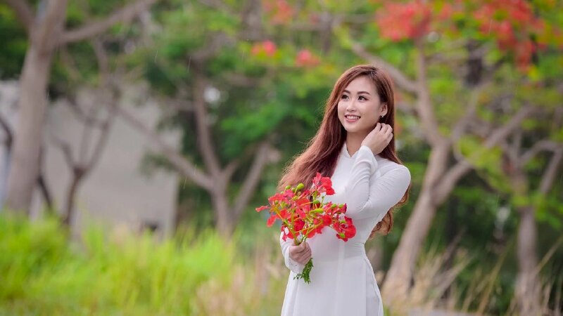 Tạo dáng đứng dễ thương trong chiếc áo dài trắng học sinh với tay cầm nhánh hoa phượng vỹ đỏ, tay còn lại vén tóc, miệng cười tươi, mắt nhìn lên phía xa