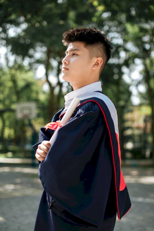 Tạo dáng nam chụp từ góc nghiêng trong bộ áo cử nhân với kiểu đứng khoanh tay, 1 tay cầm giấy cuộn tròn thể hiện sự trưởng thành và chất từ bên trong