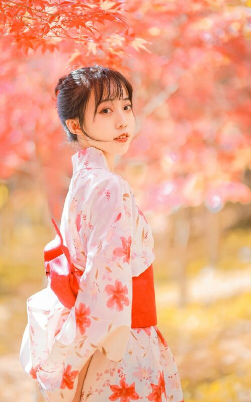 Tạo dáng chụp ảnh trong bộ áo truyền thống Nhật Bản Kimono với góc chụp nghiêng, mặt quay về nhìn ống kính, 2 tay để tạo dáng phía sau lưng dưới rừng lá phông đỏ