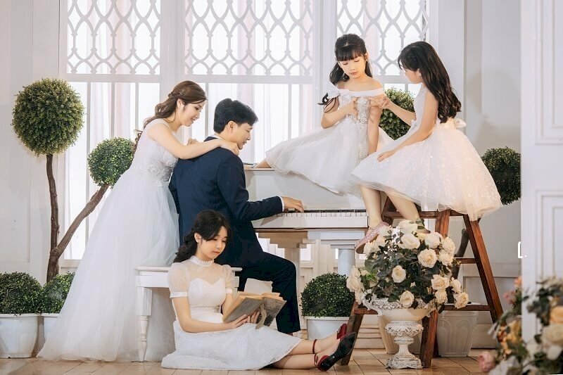Cách tạo dáng chụp ảnh gia đình 5 người kiểu tự nhiên vô cùng đáng yêu và sáng tạo khi bố ngồi đánh đàn piano, mẹ đứng sau lưng quàng tay lên vai bố, hai cô con gái ngồi trên đàn đang chơi đùa với nhau và chị gái lớn ngồi dưới sàn đang đọc sách tựa vào ghế bố.