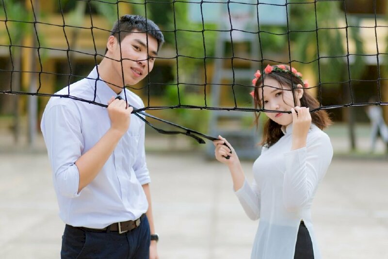 Cách tạo dáng chụp ảnh kỷ yếu đôi nam nữ cute khi đứng cầm lưới bóng chuyền, bạn nam đưa 1 tay cầm lên lưới bóng chuyền và 1 tay thả dọc theo thân người, bạn nữ đưa 1 tay cầm lên lưới bóng chuyền còn 1 tay cầm cà vạt nam và kéo lại phía mình.