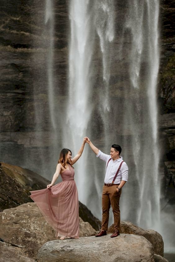 Cách tạo dáng chụp ảnh với thác nước cho cặp đôi nam nữ đang đứng bên thác nước, với kiểu này nam đứng cho 1 tay vào túi quần, 1 tay cầm tay bạn nữ đưa lên cao, nữ đứng 1 tay cầm váy xoè ra, 1 tay cầm tay bạn nam như đang múa.