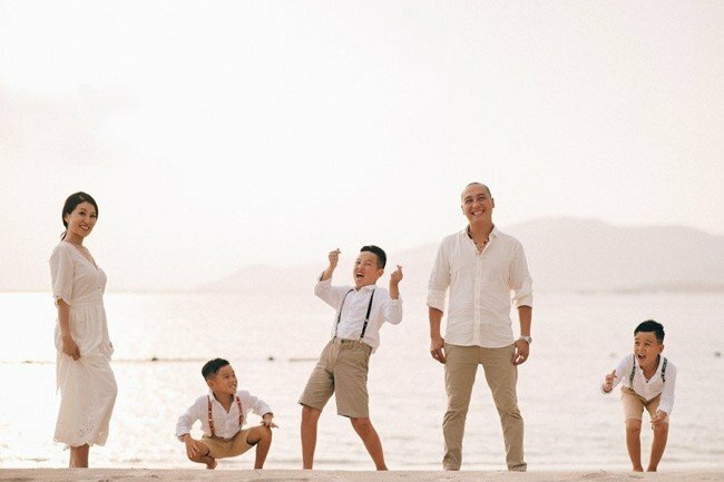 Cách tạo dáng chụp ảnh gia đình 5 người kiểu tự nhiên trên biển với kiểu 3 người cùng xếp vào 1 hàng ngang, bố đứng thẳng nhìn vào máy ảnh, bên trái bố là 1 cậu con trai, bên phải bố là 2 cậu con trai và mẹ đứng nghiêng người ngoài cùng, cả nhà cùng tươi cười vui vẻ, 3 cậu con trai thì đang đùa nghịch.