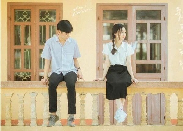 Cách tạo dáng chụp ảnh kỷ yếu đôi nam nữ học sinh với nam mặc quần tây áo sơ mi trắng và nữ mặc váy đen áo sơ mi trắng, cùng ngồi trên thành lang cang thả chân xuống, nữ để 2 bàn chân chéo lên nhau, với hai tay đặt trên lang cang.