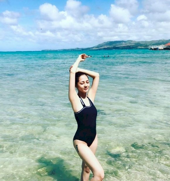 Cách tạo dáng chụp ảnh bikini ở biển với dáng đứng, 2 tay đưa thẳng qua đầu, 1 tay vòng lấy cầm lấy cánh tay kia, chân đứng 1 chân thẳng, 1 chân còn lại hơi co lên.