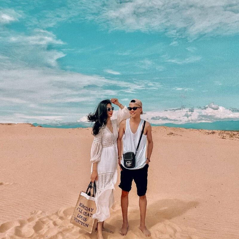 Cách tạo dáng chụp ảnh trên đồi cát cho 1 nam và 1 nữ, hai người đứng cạnh nhau, bạn nữ đặt cánh tay lên vai bạn nam và đưa tay lên đầu, bạn nữ đứng với 1 chân trước và 1 chân sau, bạn nam đứng 2 chân rộng bằng vai và cho tay vào túi quần.