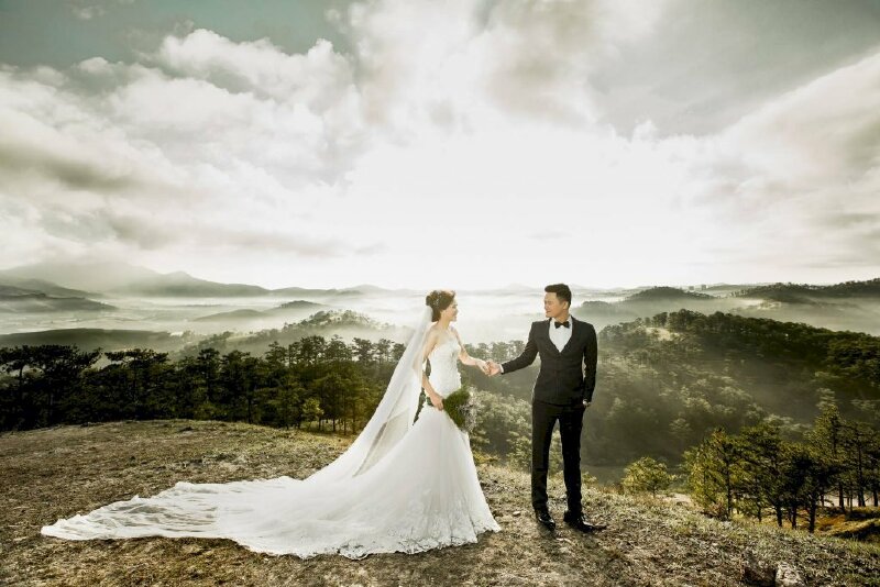 Cách tạo dáng chụp ảnh cưới ngoại cảnh ở trên đồi thông sáng sớm, ở xa xa là những  màn sương mù và ánh mặt trời đang dần lên, chú rể đứng trước cầm lấy tay cô dâu. 