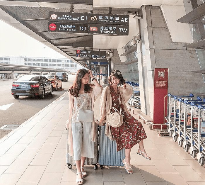 Cách tạo dáng chụp ảnh ở sân bay cho hai bạn nữ cực kỳ dễ thương khi đứng ngồi cạnh vali với một bạn đứng tựa người và đặt tay lên vali còn một tay đưa lên đầu, đứng một chân trước và một chân sau; còn bạn kia ngồi lên vali đặt một chân chạm xuống đất, một tay chống lên vali, một tay đưa lên đầu.