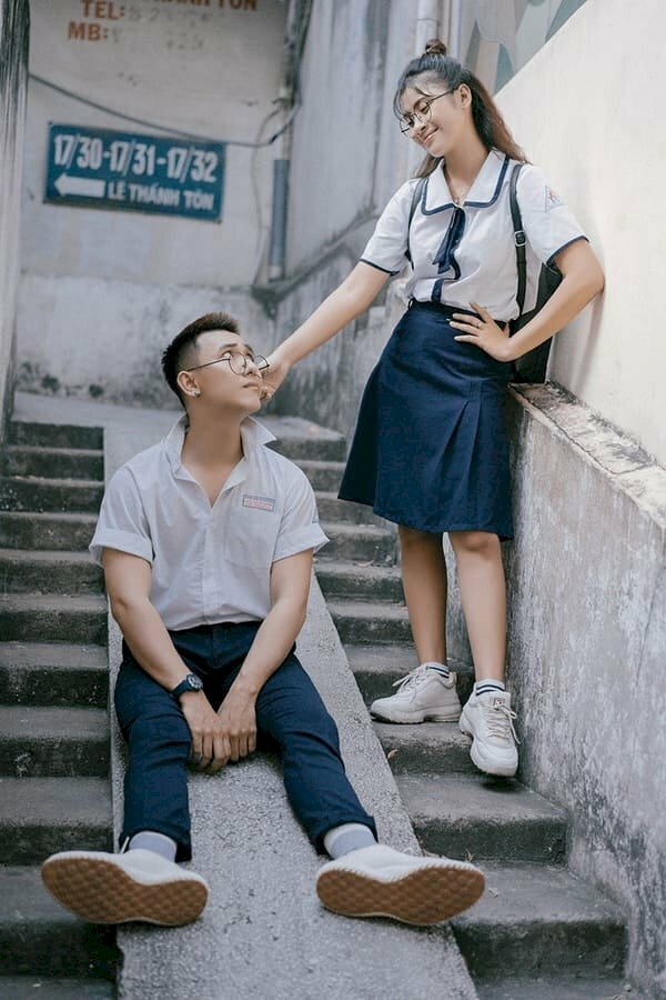 Cách tạo dáng chụp ảnh kỷ yếu đôi nam nữ trong trang phục học sinh vô cùng đáng yêu ở bậc thang lên xuống, nam ngồi giữ bậc thang để hai chân duỗi thẳng, mắt ngước nhìn nữ, bạn nữ đứng trên bậc thang với 1 tay chống nạnh và tay còn lại xách tai bạn nam. 