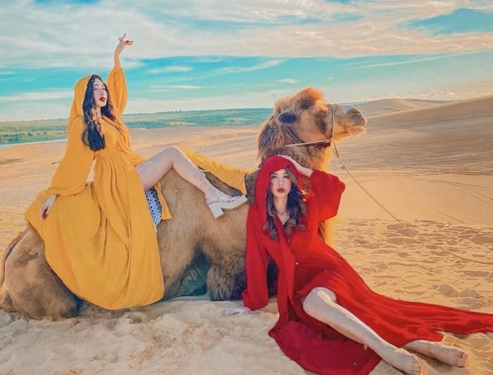 Cách tạo dáng chụp ảnh trên đồi cát cho đôi bạn thân nữ như ở Dubai vô cùng sang chảnh, một bạn nữ mặc váy dài màu vàng, 1 bạn mặc váy dài đỏ, ngồi chụp cạnh con lạc đà, 1 bạn ngồi trên lưng lạc đà tạo dáng ưỡn người ra sau đưa 1 tay thẳng ra sau và 1 tay chỉ lên trời, chân đưa lên phía trước vuông góc; bạn nữ ngồi trên cát, bắt chéo chân này qua chân kia, 1 tay chống xuống cát và 1 tay đưa lên giữ nón.