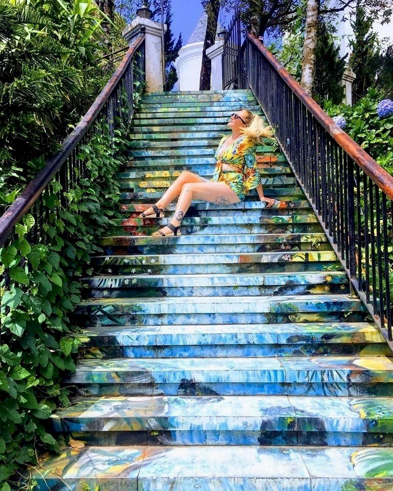 Cách tạo dáng chụp ảnh ở bậc thang con đường bích họa bằng cách ngồi nghiêng người trên bậc thang, đặt 1 chân trước, 1 chân sau, đưa tay chống sau lưng, mặt hơi ngước lên nhìn trời. 