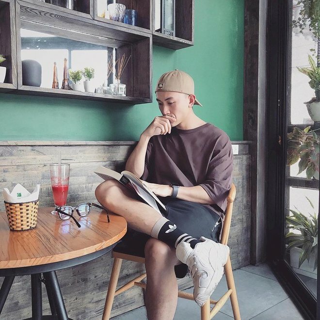 Cách tạo dáng chụp ảnh nam ngồi trong quán cafe cực ngầu với chân bắt lên hình chữ ngũ, 1 tay lấy che miệng, 1 tay đang cầm quyển sách đặt trên chân.