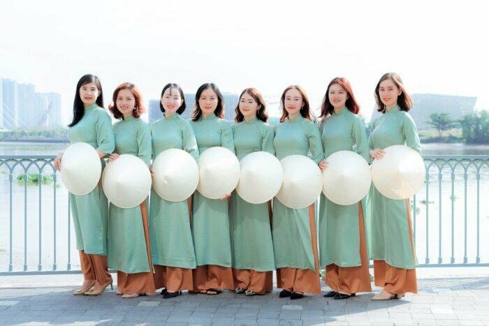 Cách tạo dáng chụp ảnh áo dài tập thể kiểu nhẹ nhàng với nhóm nữ 8 người, đạo cụ chụp là chiếc nón lá, mọi người đứng xếp thành hàng ngang, đứng nép vào nhau, tay cầm nón lá đặt ngay bụng.