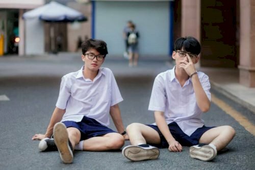 Tạo dáng chụp ảnh kỷ yếu đôi nam học sinh cấp 2 với dáng ngồi bệt xuống sân trường, 1 bạn đưa tay chống sau lưng với 1 chân duỗi thẳng, 1 chân co chữ ngũ, còn bạn kia đưa 1 tay lên đẩy kính. 