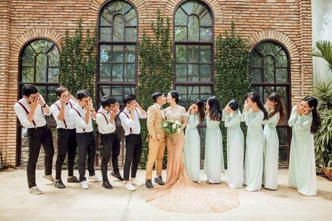 Cách tạo dáng đẹp khi đi đám cưới cùng cô dâu chú rể với kiểu xếp thành 1 hàng ngang nữ đứng bên phía cô dâu và nam đứng bên phía chú rể, mọi người cùng nhìn về phía cô dâu chú rể đưa tay lên che mắt nhưng vẫn nhìn lén họ đang hôn nhau. 