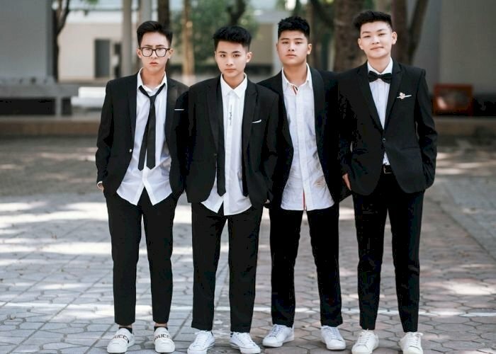 Cách tạo dáng chụp ảnh kỷ yếu cho nam thường gặp với bốn bạn học sinh diện áo vest đen mắt, đứng thẳng tự nhiên mắt nhìn ống kính và tay bỏ vào túi quần tạo sự lịch lãm, trưởng thành