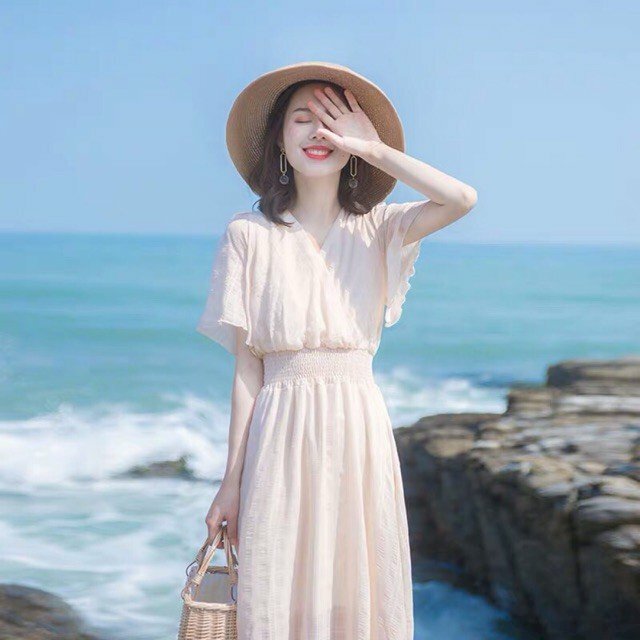 Cách tạo dáng chụp ảnh đi biển dành cho bạn nữ mặc váy dài maxi với góc nhìn đứng quay lưng lại với biển, đứng thẳng người, 1 tay cầm túi xách, 1 tay đưa lên che 1 bên mắt với bàn tay ngửa ra ngoài, miệng cười tươi, mắt nhắm và đầu hơi ngẩng lên.