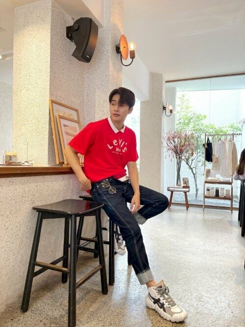 Kiểu dáng ngồi nam trong quán cafe với chiếc áo thun đỏ, quần jean vào đôi sneaker năng động, chân co gác lên ghế, chân còn lại gũi thẳng, 1 tay đặt túi quần, 1 tay đặt nhẹ lên đùi, mặt nhìn nghiêng