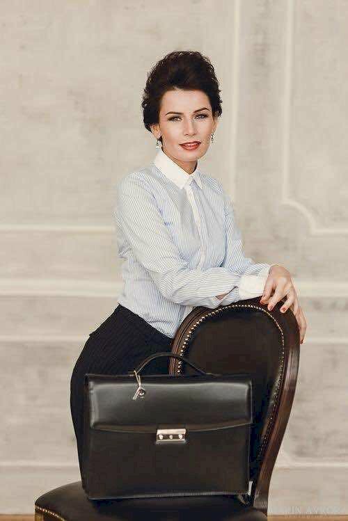 Kiểu tạo dáng quý bà doanh nhân trong bộ quần áo công sở bên chiếc ghế gỗ với kiểu đứng tựa vào ghế, 2 tay đặt lên vành ghế, miệng cười nhẹ