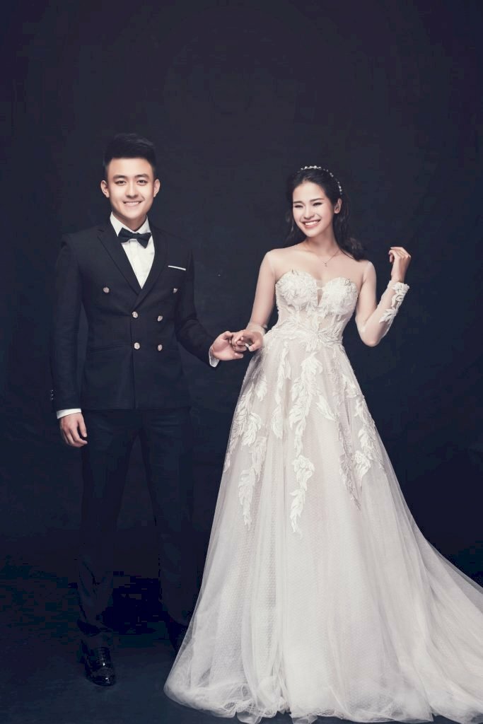 Cách tạo dáng chụp ảnh cưới trong studio theo phong cách Hàn Quốc bằng cách chú rể đững thẳng người, giang chân rộng bằng vai, cầm lấy tay cô dâu và cô dâu đứng nghiêng hông sang bên phía chú rể với gương mặt cười tươi.