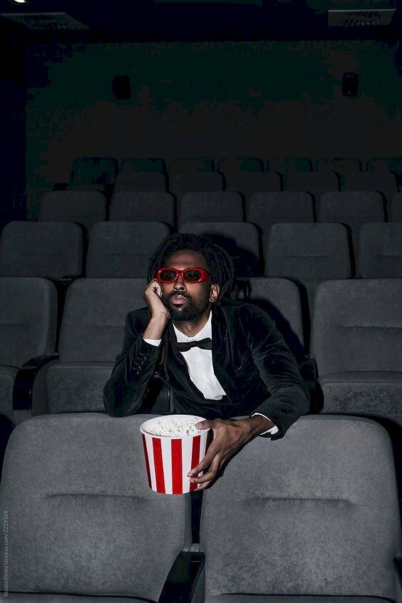 Tạo dáng quý ông lịch lãm trong phòng chiếu phim với bộ vest đen ngồi một tay chống càm, tay cầm ly bắp giữa hàng ghế đen