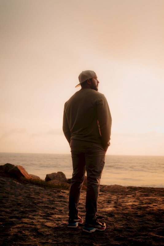 Tạo dáng chụp với cảnh biển chill phết với góc chụp từ phía sau lưng, đầu đội mũ lưỡi trai ngược, mặt nhìn nghiêng, 2 tay đặt túi quần ở phía trước, mặt hướng về biển xanh dưới hoàng hôn