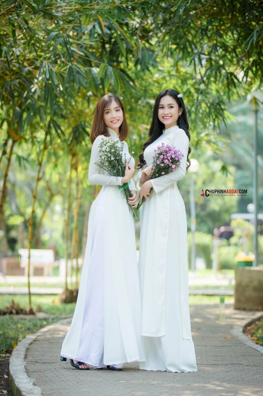 Tạo dáng dễ thương cho 2 bạn nữ khi chụp kỷ yếu trong bộ áo dài trắng người hơi nghiêng vào nhau so với ống kinh, tay cầm bó hoa, miệng cười tươi