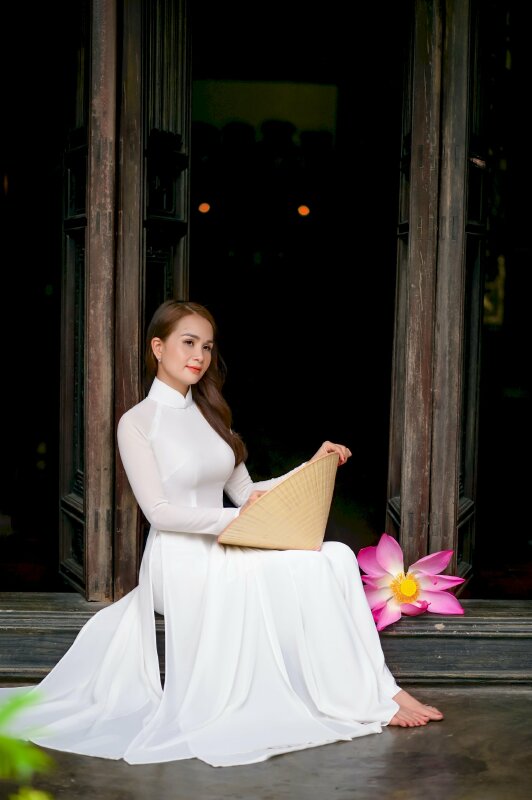 Tạo dáng đẹp trong chiếc áo dài trắng với tư thế ngồi tựa lưng vào cột người hơi nghiêng, 2 tay với cầm nón lá đặt trên đùi, tóc xõa một bên, đầu hơi nghiêng, mắt nhìn xa