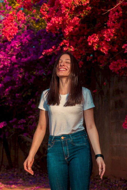 Tạo dáng chụp ảnh ngoại cảnh với kiểu đứng tự nhiên bên khóm hoa đỏ, 2 tay xuôi thẳng, đầu ngước lên, mắt nhắm, miệng cười tươi