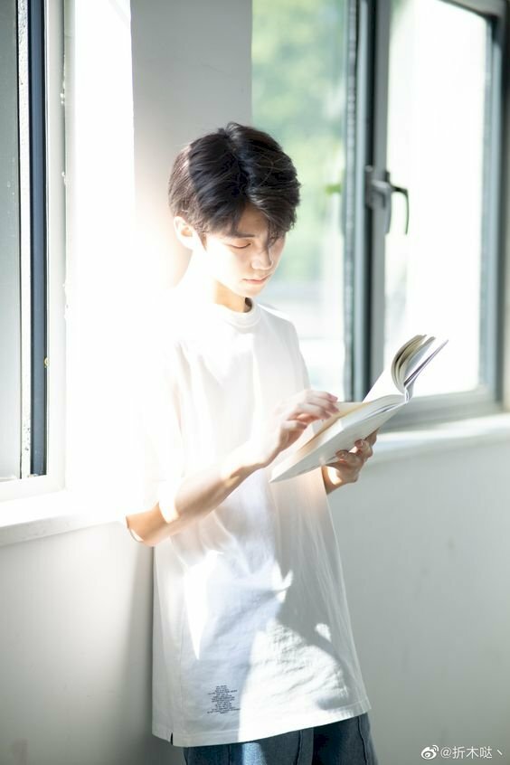 Cách tạo dáng chụp ảnh cho học sinh nam với dáng đứng thẳng dựa vào tường, 1 tay cầm quyển sách, 1 tay đang lật trang sách, mặt hơi cúi nhìn vào sách.