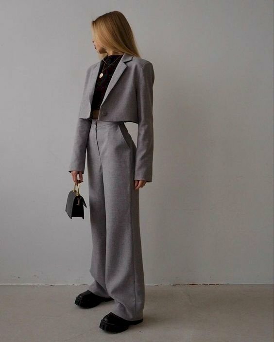 Cách tạo dáng chụp ảnh với vest nữ khi đang đứng nghiêng người với vai hơi nghiêng về phía ống kính, đầu quay đi, 1 tay để thẳng, 1 tay cầm túi xách để thẳng trên đùi.