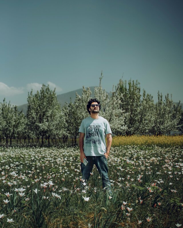 Cách chụp ảnh giữa cánh đồng hoa với dáng đứng tự nhiên thoải mái đầu hơi ngẩng lên mắt mang kính đen, một tay cầm điện thoại, 1 tay đặt túi quần