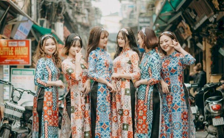 Cách tạo dáng chụp ảnh áo dài tập thể kiểu tự nhiên dành cho nhóm bạn 5 người, với áo dài màu sắc có họa tiết tương đồng liên quan với nhau, cùng xếp thành 1 hàng ngang đứng hơi nghiêng người néo vào nhau, với biểu cảm tự nhiên.