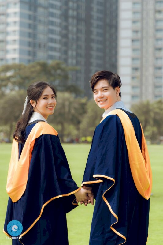 Cách tạo dáng chụp ảnh áo cử nhân kỷ yếu đôi nam nữ đang nắm tay nhau bước đi và cùng quay đầu nhìn lại cười rất tươi.