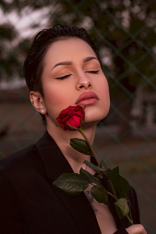Cô gái trong chiếc áo đen cùng hình xăm hoa hồng trên ngực, chụp nửa người lấy góc nghiêng, mặt hơi ngước, mắt nhắm nhẹ, 1 tay cầm hoa hồng chạm má chill trong buổi chiều nắng tắt
