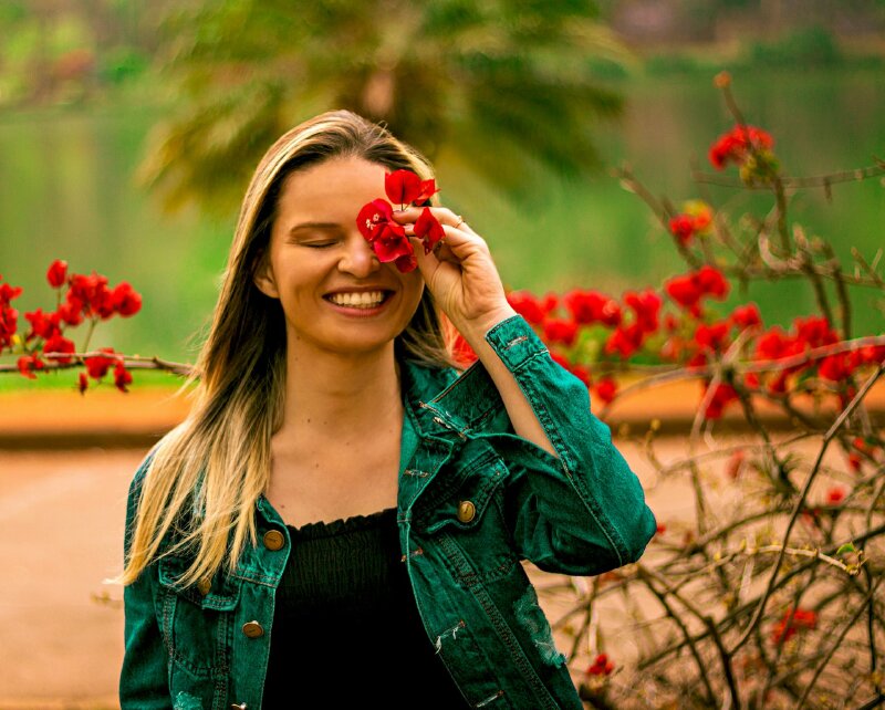 Kiểu chụp ảnh nửa người đẹp bên khóm hoa giấy màu đỏ với cách tạo dáng chụp ảnh đẹp ngoài trời đứng thẳng, một tay đưa hoa giấy che một bên mắt, 2 mắt nhắm cùng miệng nở nụ cười tươi