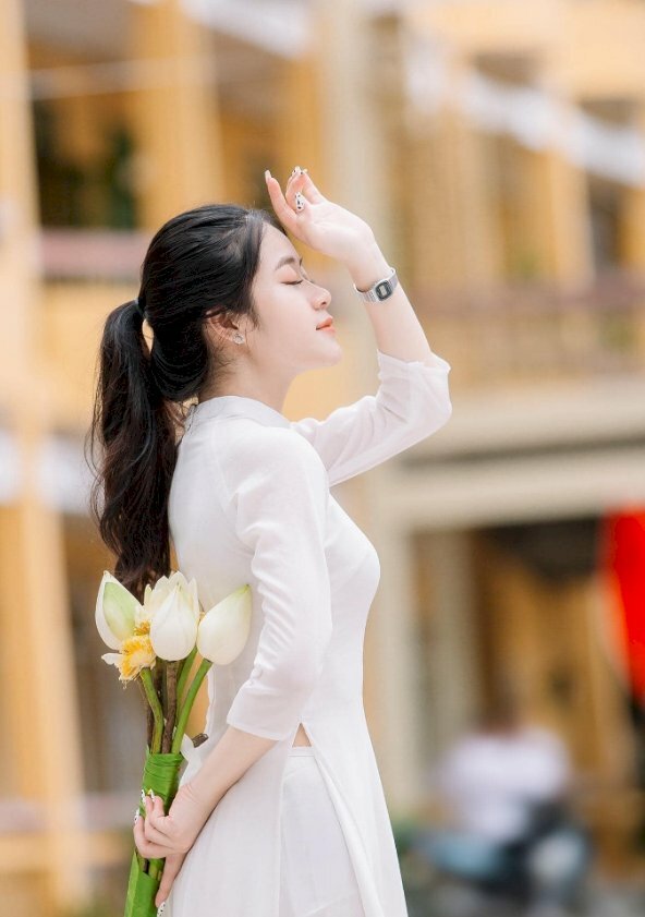 Kiểu dáng chụp kỷ yếu với áo dài trắng cực chill với một tay che trên trán 1 tay cầm bó hoa sen trắng giấu sau lưng, mắt nhắm nhẹ, mặt hơi ngước lên