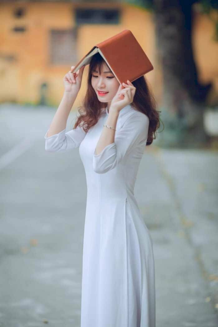 Kiểu tạo dáng nữ siêu cute với chiếc áo dài trắng và 2 tay cầm quyển sách đội che nắng, mắt nhìn xuống, miệng mỉm cười